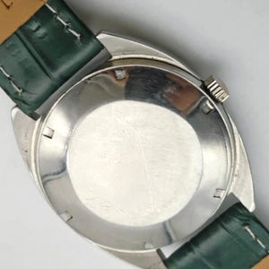Puede incluir: Primer plano de un reloj de pulsera plateado con una correa de cuero verde. La esfera del reloj no es visible. La correa est&aacute; sujeta a la caja del reloj con un respaldo de cuero marr&oacute;n claro. La corona del reloj es visible.