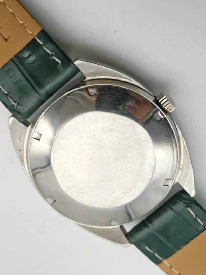Puede incluir: Primer plano de un reloj de pulsera plateado con una correa de cuero verde. La esfera del reloj no es visible. La correa est&aacute; sujeta a la caja del reloj con un respaldo de cuero marr&oacute;n claro. La corona del reloj es visible.