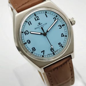 K&ouml;nnte beinhalten: Eine silberfarbene Armbanduhr mit hellblauem Zifferblatt und schwarzen Ziffern. Die Uhr hat ein braunes Lederarmband und das Jaeger-LeCoultre Club-Logo. Die Zeiger der Uhr sind schwarz und die Uhr ist mit "SWISS MADE" gekennzeichnet.