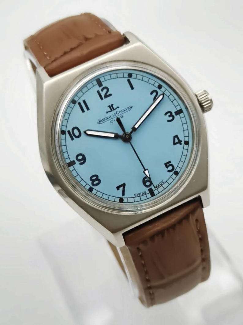 K&ouml;nnte beinhalten: Eine silberfarbene Armbanduhr mit hellblauem Zifferblatt und schwarzen Ziffern. Die Uhr hat ein braunes Lederarmband und das Jaeger-LeCoultre Club-Logo. Die Zeiger der Uhr sind schwarz und die Uhr ist mit "SWISS MADE" gekennzeichnet.