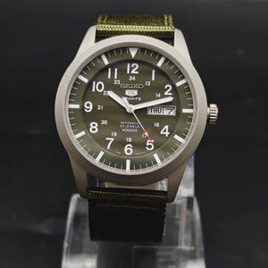 Puede incluir: Un reloj de pulsera autom&aacute;tico Seiko 5 Sports con esfera verde oliva, n&uacute;meros ar&aacute;bigos blancos y una ventana de d&iacute;a y fecha en la posici&oacute;n de las tres. El reloj cuenta con una caja plateada y una correa de tela verde oliva a juego.