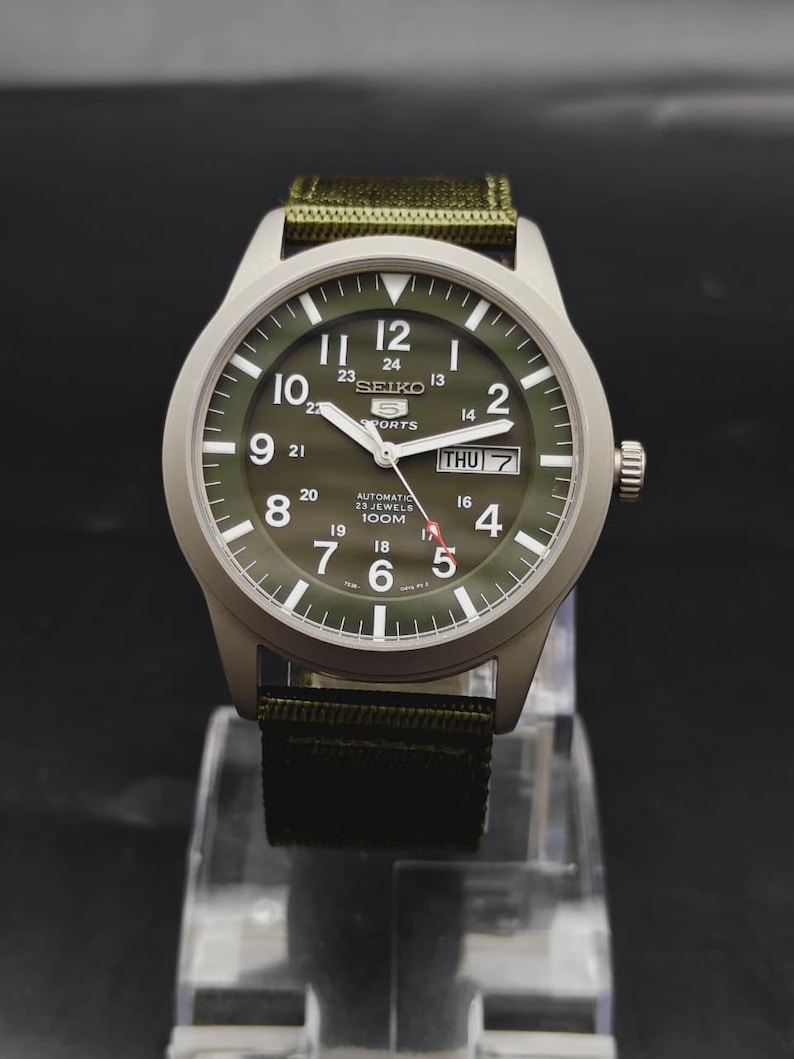 Puede incluir: Un reloj de pulsera autom&aacute;tico Seiko 5 Sports con esfera verde oliva, n&uacute;meros ar&aacute;bigos blancos y una ventana de d&iacute;a y fecha en la posici&oacute;n de las tres. El reloj cuenta con una caja plateada y una correa de tela verde oliva a juego.
