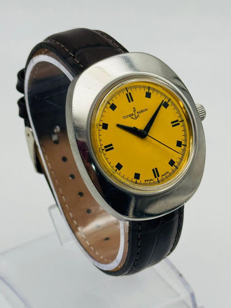 May include: A vintage Ulysse Nardin wristwatch with a yellow face and black hands. The watch has a silver-coloured case and a dark brown leather strap. The watch face displays the brand name and is marked "Swiss Made".