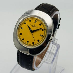 May include: A vintage Ulysse Nardin wristwatch with a silver case and a bright yellow face. The watch features black hands and hour markers, and a dark brown leather strap. The watch is displayed on a clear stand.