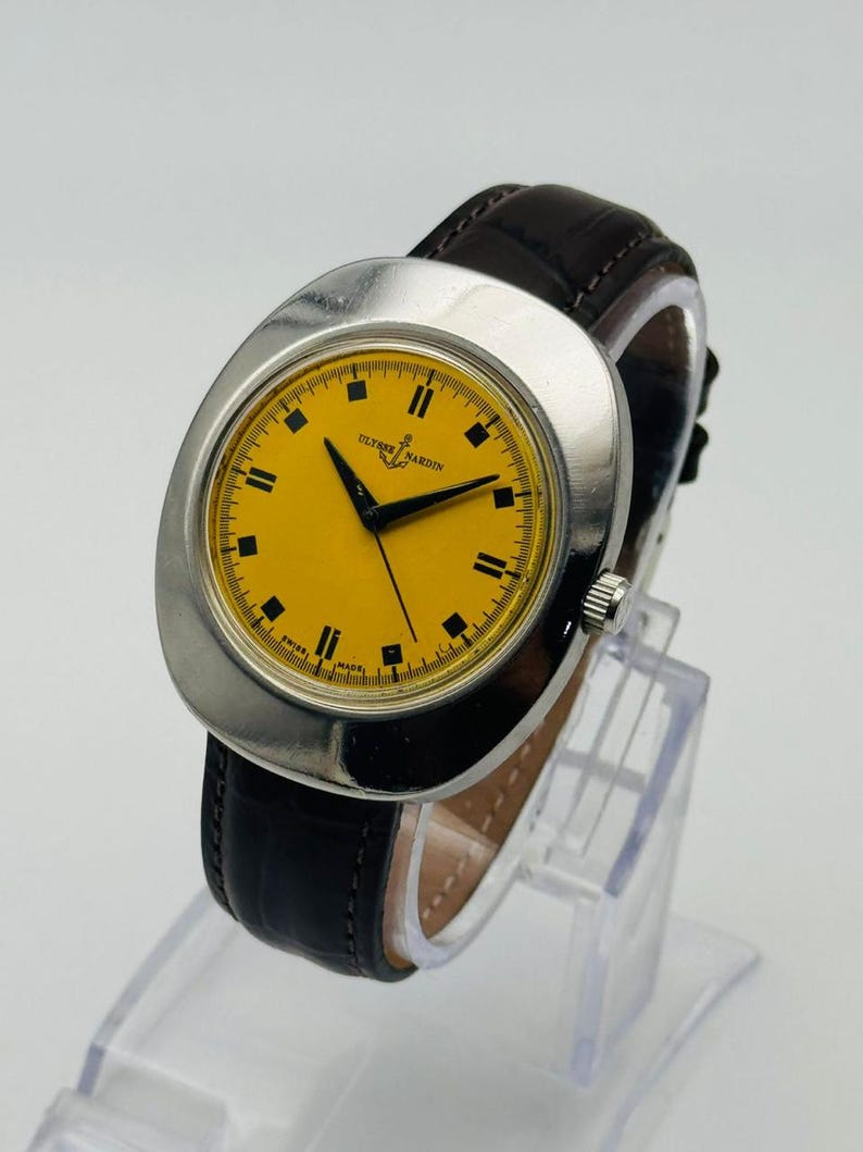 May include: A vintage Ulysse Nardin wristwatch with a silver case and a bright yellow face. The watch features black hands and hour markers, and a dark brown leather strap. The watch is displayed on a clear stand.