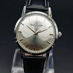 Op de afbeelding: Een zilverkleurig Jaeger-LeCoultre Club horloge met een zwarte leren band. De wijzerplaat heeft een geborstelde afwerking en is voorzien van de merknaam en de tekst "Swiss Made". Het horloge wordt gepresenteerd op een transparante standaard tegen een donkere achtergrond.