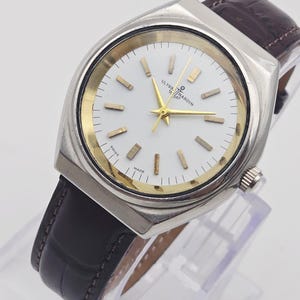 Pu&ograve; includere: Un orologio Ulysse Nardin argentato con quadrante bianco, dettagli dorati e cinturino in pelle marrone. L'orologio ha una cassa rettangolare e un design classico. Il quadrante mostra il testo "Ulysse Nardin" e "Swiss Made".