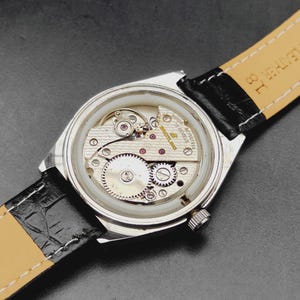 May include: A silver-toned wristwatch with the back casing removed to reveal the internal mechanical movement. The movement features gold-toned plates, gears, and the text Breitling 17 Jewels. It is attached to a black leather strap with a tan lining.