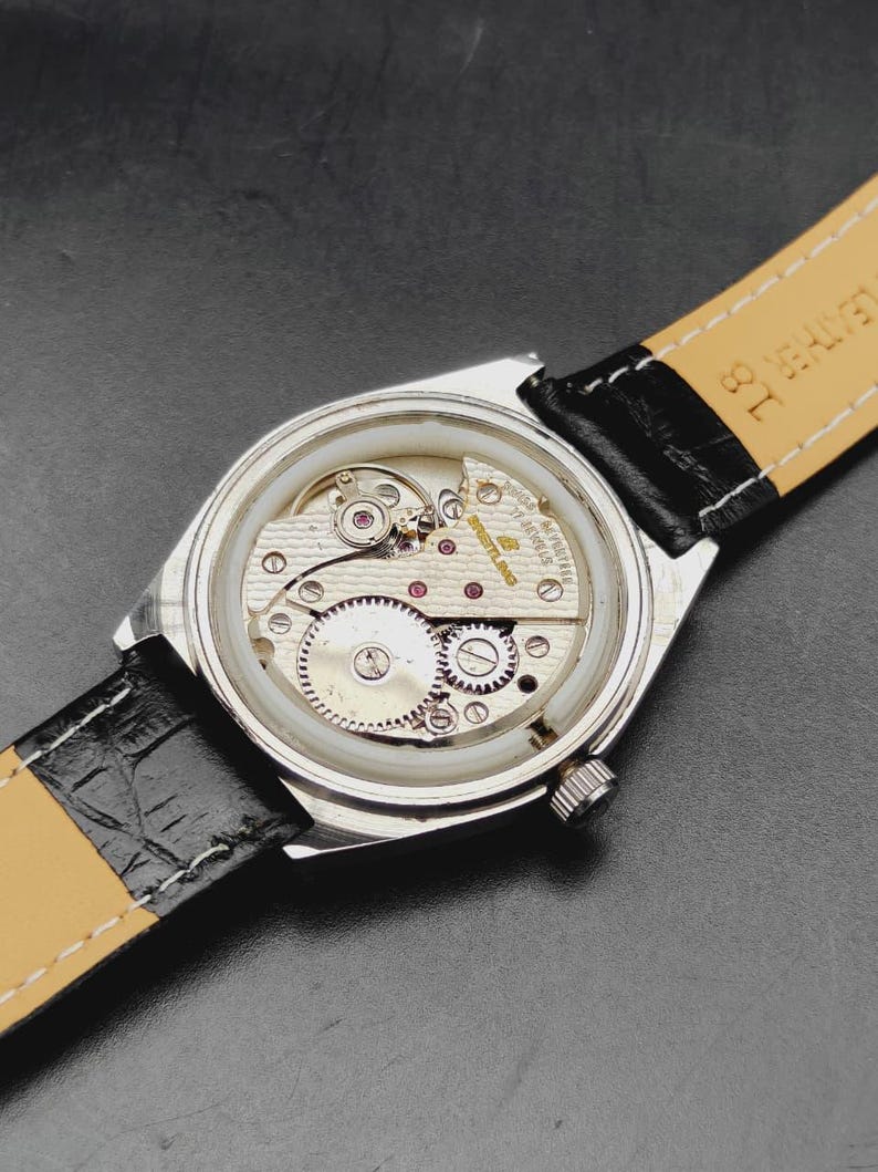 May include: A silver-toned wristwatch with the back casing removed to reveal the internal mechanical movement. The movement features gold-toned plates, gears, and the text Breitling 17 Jewels. It is attached to a black leather strap with a tan lining.