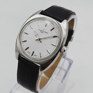 May include: A silver-toned Ulysse Nardin watch with a white face and black leather band. The watch has a rectangular case and a crown on the right side. The watch face features the brand name and simple hour markers.