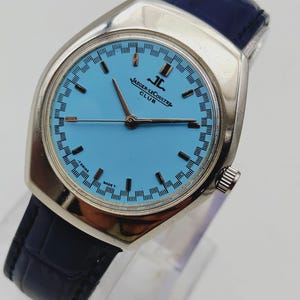 May include: A vintage Jaeger-LeCoultre Club watch with a light blue dial and silver case. The watch has a dark blue leather strap. The dial displays the Jaeger-LeCoultre logo and Swiss Made text.