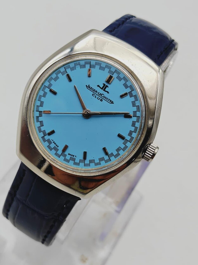 May include: A vintage Jaeger-LeCoultre Club watch with a light blue dial and silver case. The watch has a dark blue leather strap. The dial displays the Jaeger-LeCoultre logo and Swiss Made text.