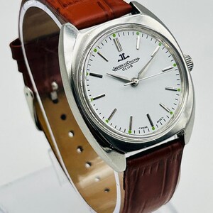 May include: A silver-toned Jaeger-LeCoultre Club watch with a white face and brown leather band. The watch face has black hour markers and green accents. The band is a rich brown color.