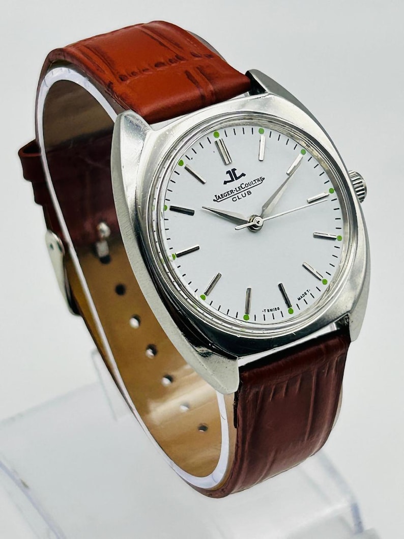 May include: A silver-toned Jaeger-LeCoultre Club watch with a white face and brown leather band. The watch face has black hour markers and green accents. The band is a rich brown color.