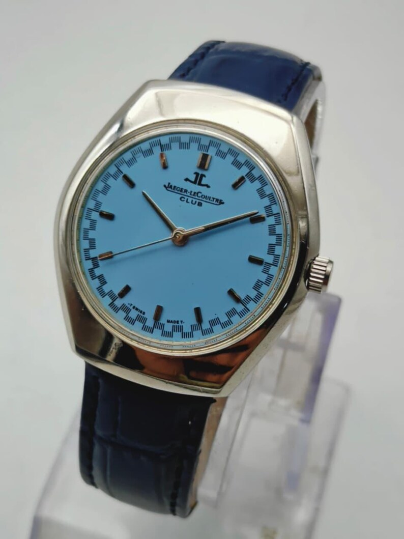 May include: A vintage Jaeger-LeCoultre Club watch with a light blue dial and silver case. The watch has a blue leather strap. The dial displays the brand name. The watch has a classic design.