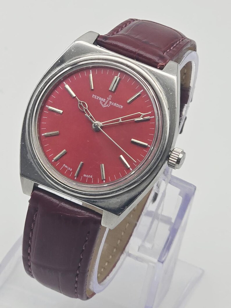 May include: A vintage Ulysse Nardin watch with a vibrant red dial and silver hands. The watch features a stainless steel case and a burgundy leather strap. The watch is displayed on a clear stand.