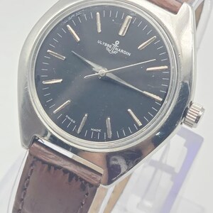May include: A vintage Ulysse Nardin wristwatch with a black dial and silver-tone hands and markers. The watch features a stainless steel case and a brown leather strap. The dial includes the brand name and the words "Swiss Made".