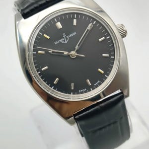 Puede incluir: Un reloj de pulsera Ulysse Nardin plateado con esfera negra y correa de cuero negro. La esfera del reloj tiene marcadores y agujas plateadas, con el nombre de la marca y el logotipo de la ancla. Est&aacute; marcado "SWISS MADE".