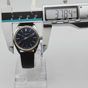 May include: A silver-toned wristwatch with a dark blue face and black leather band is measured with a digital caliper. The watch face is 37.84 mm wide. The watch is displayed on a clear plastic stand.
