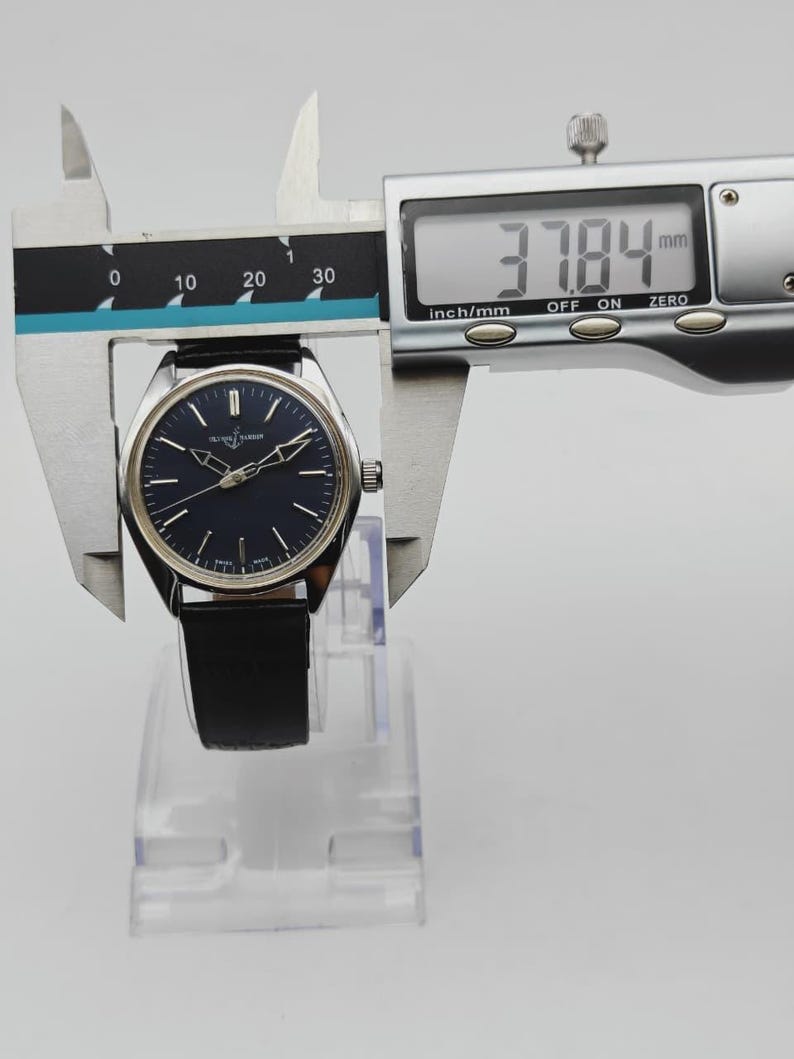 May include: A silver-toned wristwatch with a dark blue face and black leather band is measured with a digital caliper. The watch face is 37.84 mm wide. The watch is displayed on a clear plastic stand.