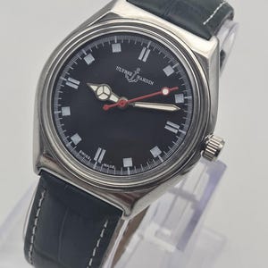 Puede incluir: Un reloj Ulysse Nardin con esfera negra y correa de cuero negra. El reloj tiene una caja plateada, marcadores de hora blancos y un segundero rojo. El reloj se exhibe sobre un soporte transparente.