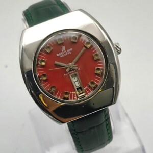 May include: A vintage Breitling Geneve automatic watch with a red face and a green leather band. The watch has a silver-colored case and a date display. The watch face features gold-colored hour markers and hands.
