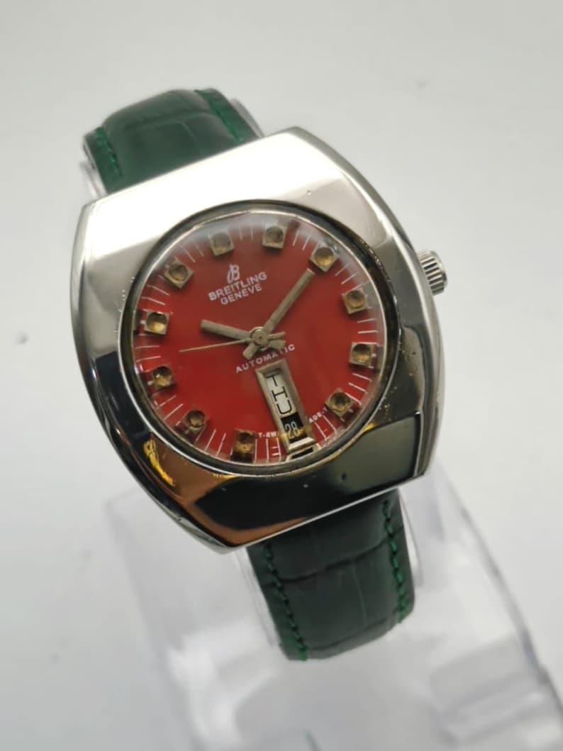 May include: A vintage Breitling Geneve automatic watch with a red face and a green leather band. The watch has a silver-colored case and a date display. The watch face features gold-colored hour markers and hands.