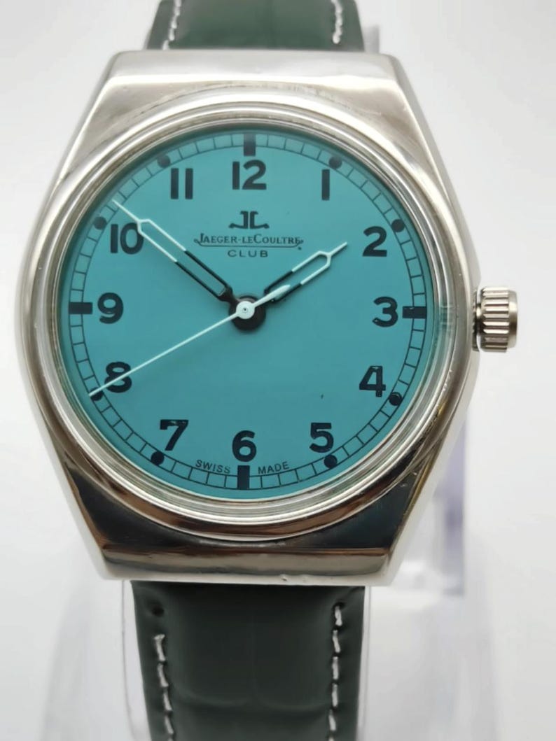 Pode incluir: Um rel&oacute;gio de pulso prateado com mostrador turquesa e algarismos pretos. O rel&oacute;gio apresenta a marca "JAEGER-LECOULTRE CLUB" e tem uma bracelete de couro verde escuro. Os ponteiros s&atilde;o prateados e o rel&oacute;gio est&aacute; marcado "SWISS MADE".