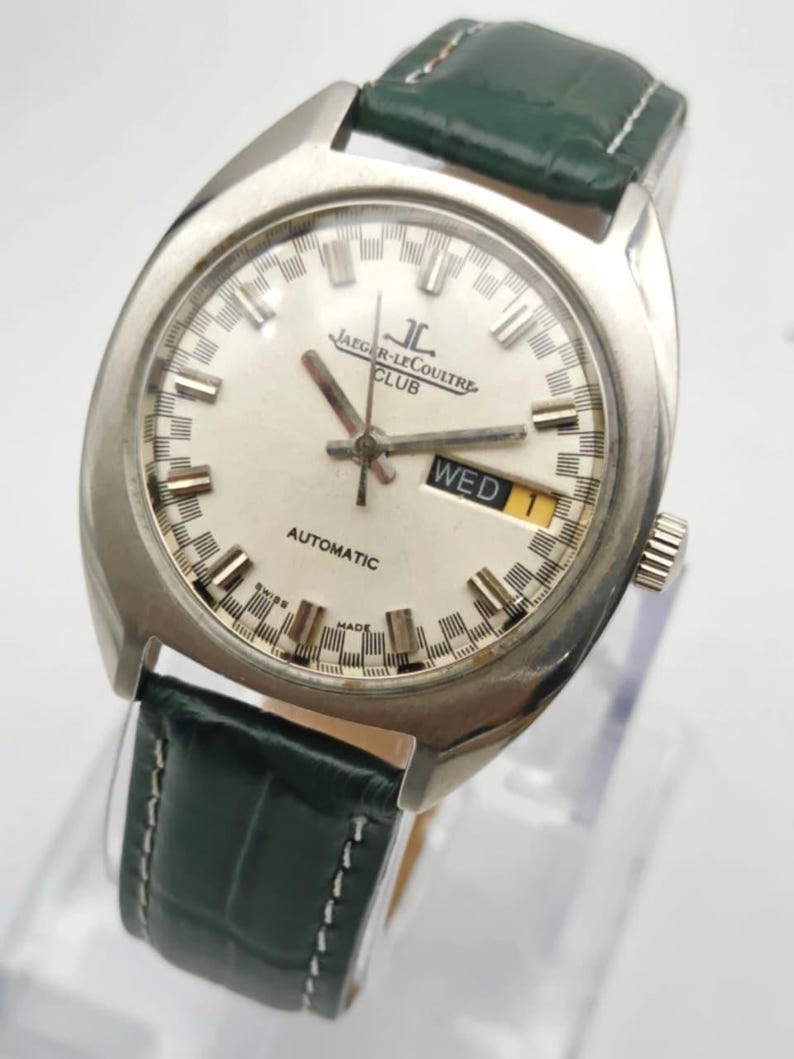 Puede incluir: Un reloj de pulsera autom&aacute;tico Jaeger-LeCoultre Club plateado con esfera blanca y correa de cuero verde. La esfera muestra el d&iacute;a de la semana y el texto "AUTOMATIC" y "MADE SWISS".