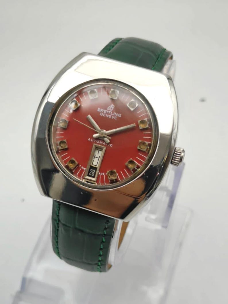 May include: A vintage Breitling Geneve automatic watch with a red dial and a date display at the 6 o'clock position. The watch has a silver-tone case and a green leather strap. The watch is displayed on a clear stand.