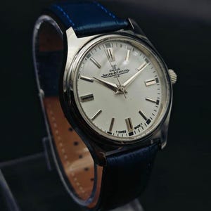 Peut inclure: Une montre Jaeger-LeCoultre Club argent&eacute;e avec un cadran blanc et des aiguilles et index argent&eacute;s. La montre a un bracelet en cuir bleu. Le cadran affiche le logo Jaeger-LeCoultre et les mots "CLUB" et "T SWISS MADE T".