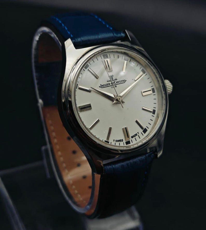 Peut inclure: Une montre Jaeger-LeCoultre Club argent&eacute;e avec un cadran blanc et des aiguilles et index argent&eacute;s. La montre a un bracelet en cuir bleu. Le cadran affiche le logo Jaeger-LeCoultre et les mots "CLUB" et "T SWISS MADE T".