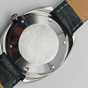 May include: A silver-toned wristwatch with a black leather strap. The watch has a round, brushed metal back and a textured crown. The strap is dark green on the outside and tan on the inside.