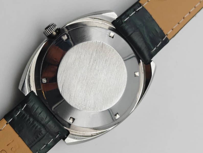 May include: A silver-toned wristwatch with a black leather strap. The watch has a round, brushed metal back and a textured crown. The strap is dark green on the outside and tan on the inside.