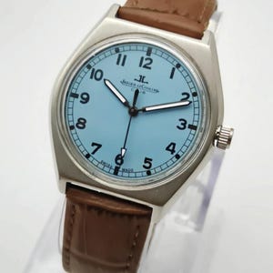 K&ouml;nnte beinhalten: Eine silberfarbene Armbanduhr mit hellblauem Zifferblatt und braunem Lederarmband. Das Zifferblatt zeigt schwarze Ziffern und Zeiger, mit dem Markennamen "Jaeger-LeCoultre". Die Uhr ist Swiss Made.