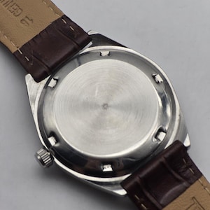 May include: Close-up of the back of a silver-toned wristwatch with a brown leather band. The watch face is circular with a brushed metal finish. The band is a dark brown color and appears to be genuine leather.