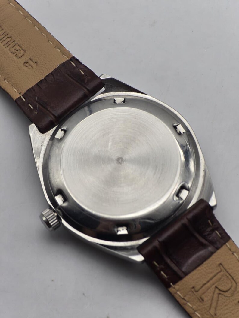 May include: Close-up of the back of a silver-toned wristwatch with a brown leather band. The watch face is circular with a brushed metal finish. The band is a dark brown color and appears to be genuine leather.