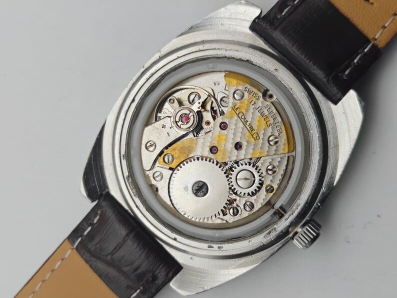 Op de afbeelding: Close-up van een vintage polshorloge met een zilverkleurige kast en een zwarte leren band. Het ingewikkelde interne mechanisme van het horloge is zichtbaar, met tandwielen, juwelen en de tekst "SWISS SEVENTEEN 17 JEWELS LE COULTRE CO."