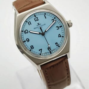 K&ouml;nnte beinhalten: Eine silberfarbene Armbanduhr mit hellblauem Zifferblatt und schwarzen Ziffern. Die Uhr hat ein braunes Lederarmband und der Text "JAEGER-LECOULTRE CLUB SWISS MADE" ist auf dem Zifferblatt zu sehen. Eine klassische Uhr.