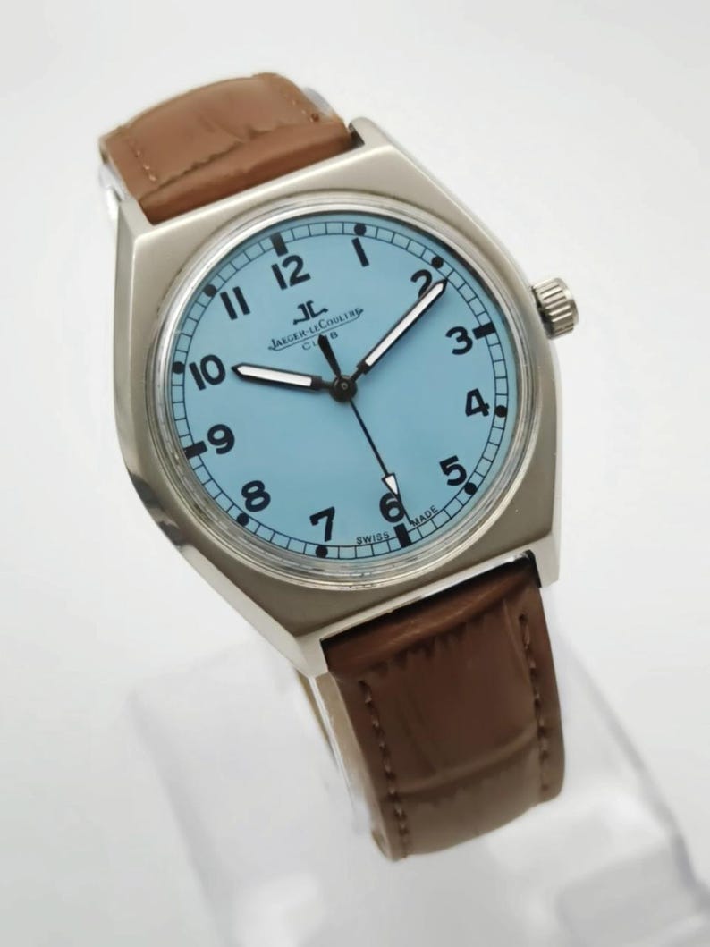 K&ouml;nnte beinhalten: Eine silberfarbene Armbanduhr mit hellblauem Zifferblatt und schwarzen Ziffern. Die Uhr hat ein braunes Lederarmband und der Text "JAEGER-LECOULTRE CLUB SWISS MADE" ist auf dem Zifferblatt zu sehen. Eine klassische Uhr.