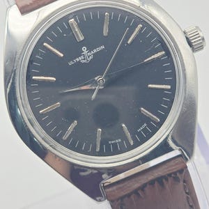 May include: A silver-toned Ulysse Nardin wristwatch with a dark face and brown leather band. The watch face features silver hour markers and hands, with the Ulysse Nardin logo and the word "SWISS" visible. The band is a dark brown color.