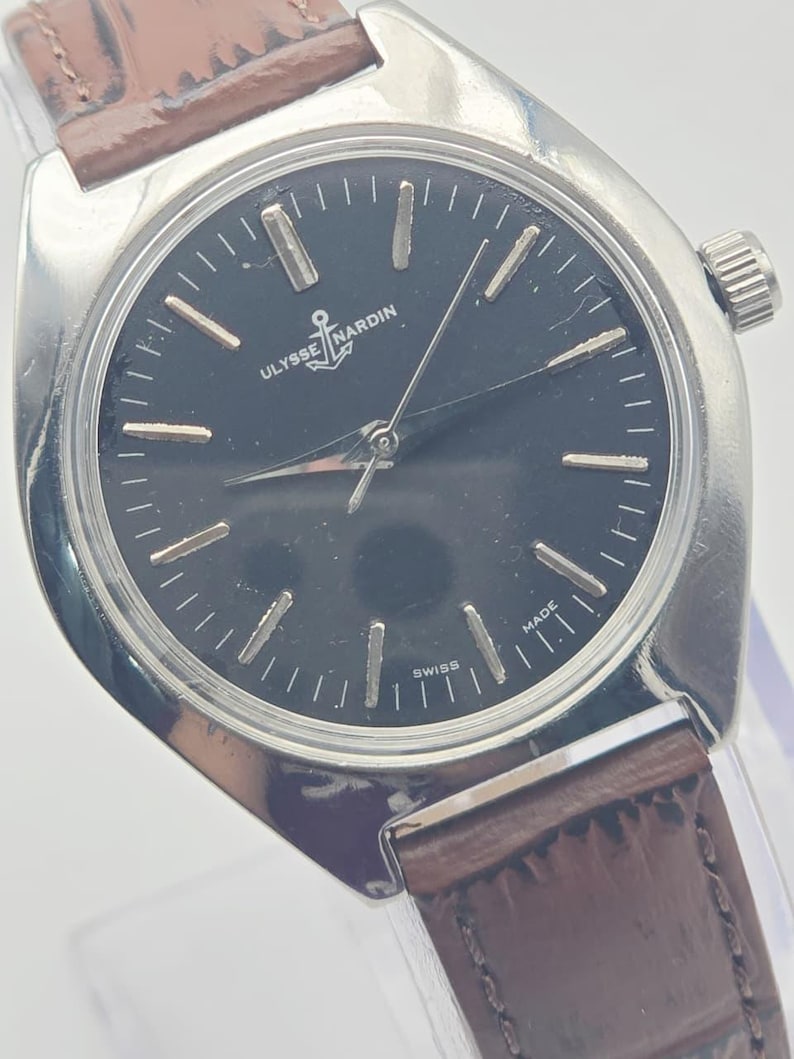 May include: A silver-toned Ulysse Nardin wristwatch with a dark face and brown leather band. The watch face features silver hour markers and hands, with the Ulysse Nardin logo and the word "SWISS" visible. The band is a dark brown color.