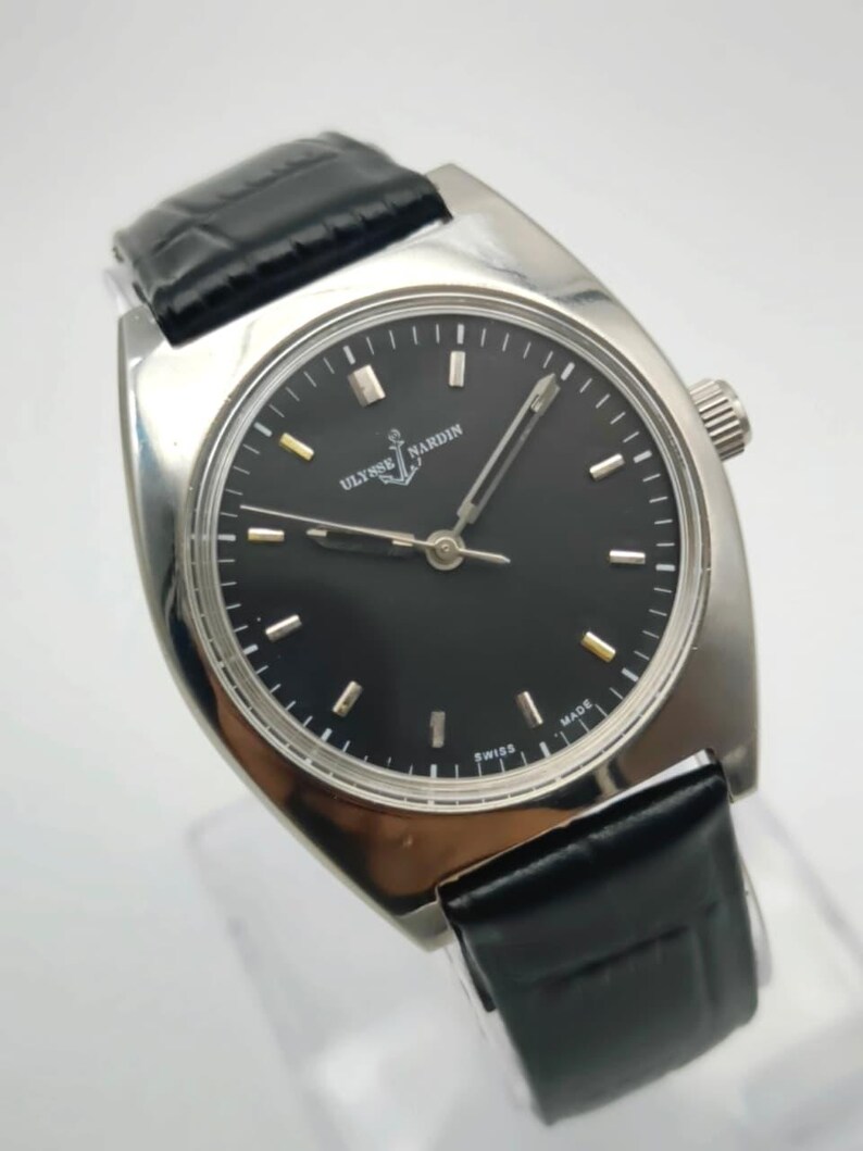 Puede incluir: Un reloj de pulsera plateado con esfera negra y correa de cuero negro. La esfera del reloj presenta marcadores de hora plateados y la marca "ULYSSE NARDIN". El reloj tiene un dise&ntilde;o cl&aacute;sico y minimalista.
