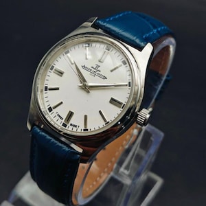 Peut inclure: Une montre Jaeger-LeCoultre Club argent&eacute;e avec un cadran blanc et un bracelet en cuir bleu. Le cadran comporte des marqueurs et des aiguilles noirs, avec le nom de la marque et le texte "SWISS MADE T". Le bracelet mesure environ 20 mm de large.