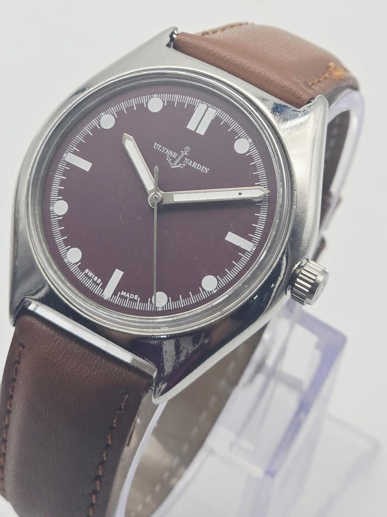 May include: A silver-toned wristwatch with a burgundy face and brown leather band. The watch face features white hour markers and hands, with the brand name "Ulysse Nardin" visible. The watch is displayed on a clear stand.