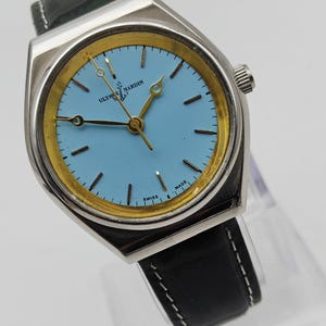 May include: A vintage Ulysse Nardin wristwatch with a light blue face and gold-tone accents. The watch has a silver-tone case and a black leather band. The watch face displays the brand name and the words "SWISS MADE".