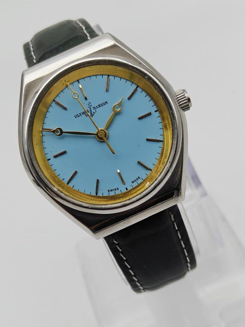 May include: A vintage Ulysse Nardin wristwatch with a light blue face and gold-tone accents. The watch has a silver-tone case and a black leather band. The watch face displays the brand name and the words "SWISS MADE".