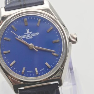May include: A silver-toned Jaeger-LeCoultre Club watch with a blue dial and a dark blue leather strap. The watch face features gold-colored hour markers and hands. The text "JAEGER-LECOULTRE CLUB" is visible on the dial. "SWISS MADE" is also printed on the watch face.