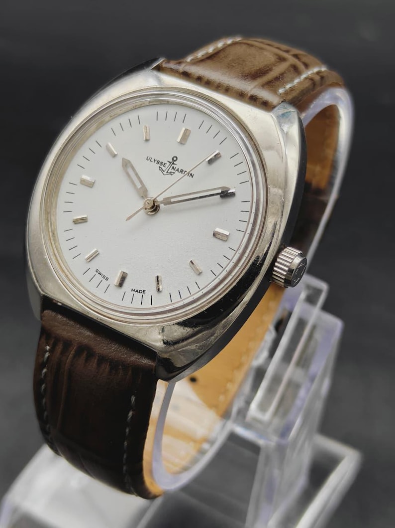 May include: A silver-tone wristwatch with a white dial, silver baton indices, and silver hands. The watch features the Ulysse Nardin logo with an anchor symbol at the twelve o'clock position and the text SWISS MADE at the bottom. It has a brown leather strap.