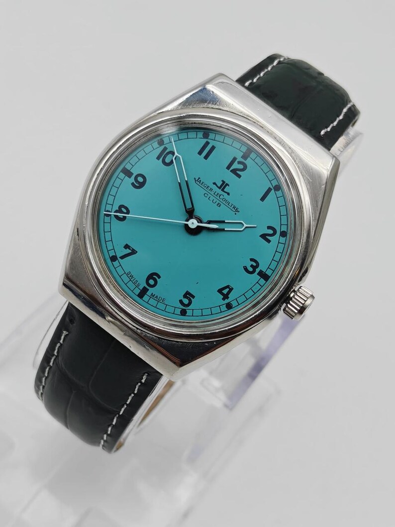 Pode incluir: Um rel&oacute;gio Jaeger-LeCoultre Club prateado com mostrador turquesa e algarismos pretos. Possui uma pulseira de couro preta com costura branca. O mostrador exibe o logotipo Jaeger-LeCoultre e "SWISS MADE".